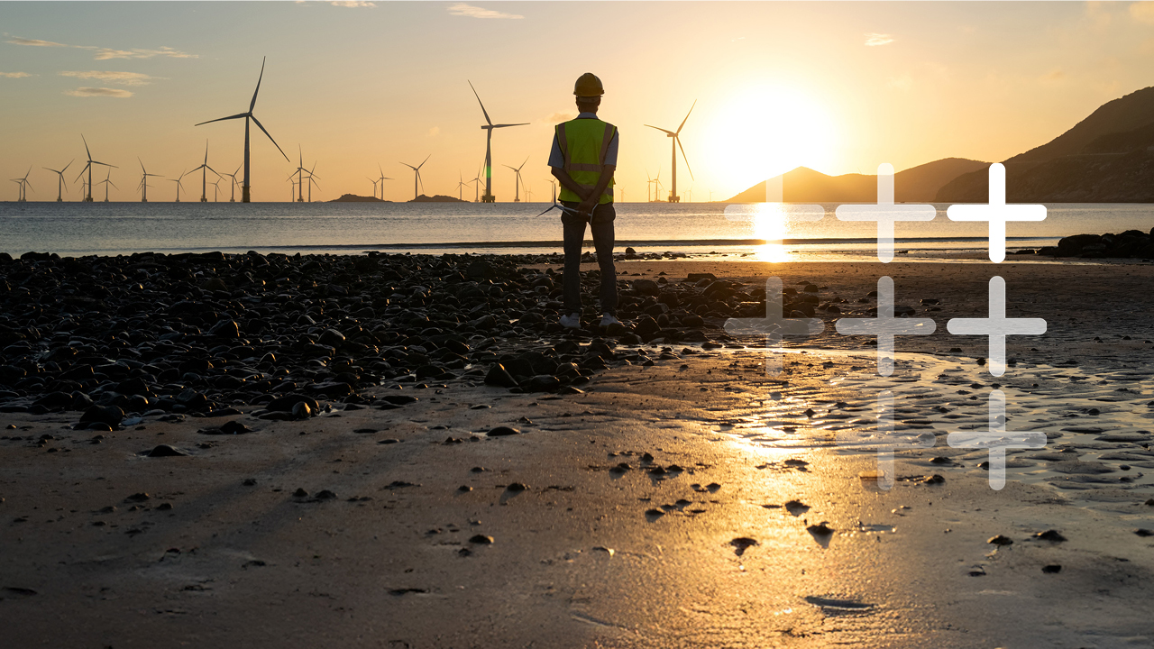 A worker wearing a hard hat standing on a beach looking out to sea with wind turbines visible in the background