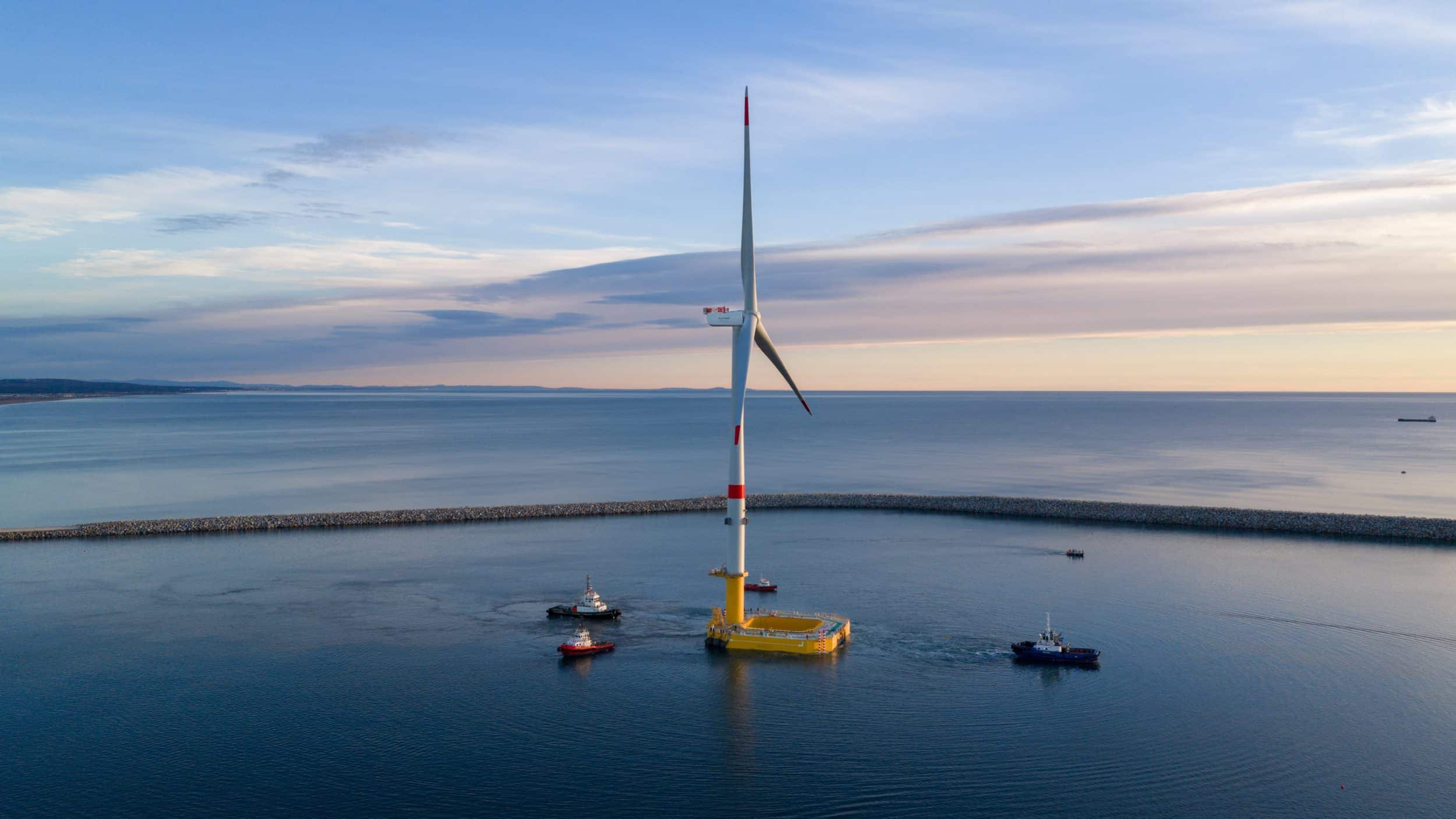 Floating offshore wind turbine being towed by multiple support vessels in calm coastal waters, showcasing floating wind installation operations and marine coordination.