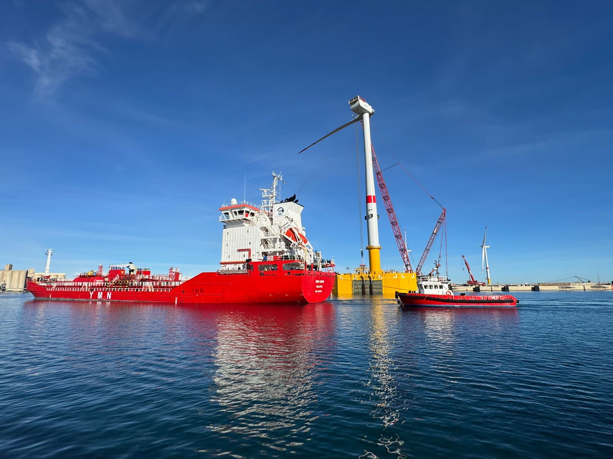 Large red offshore support vessels positioned beside a floating offshore wind turbine foundation during installation operations, with cranes and marine equipment visible at a calm port, illustrating offshore wind construction and maritime risk management.