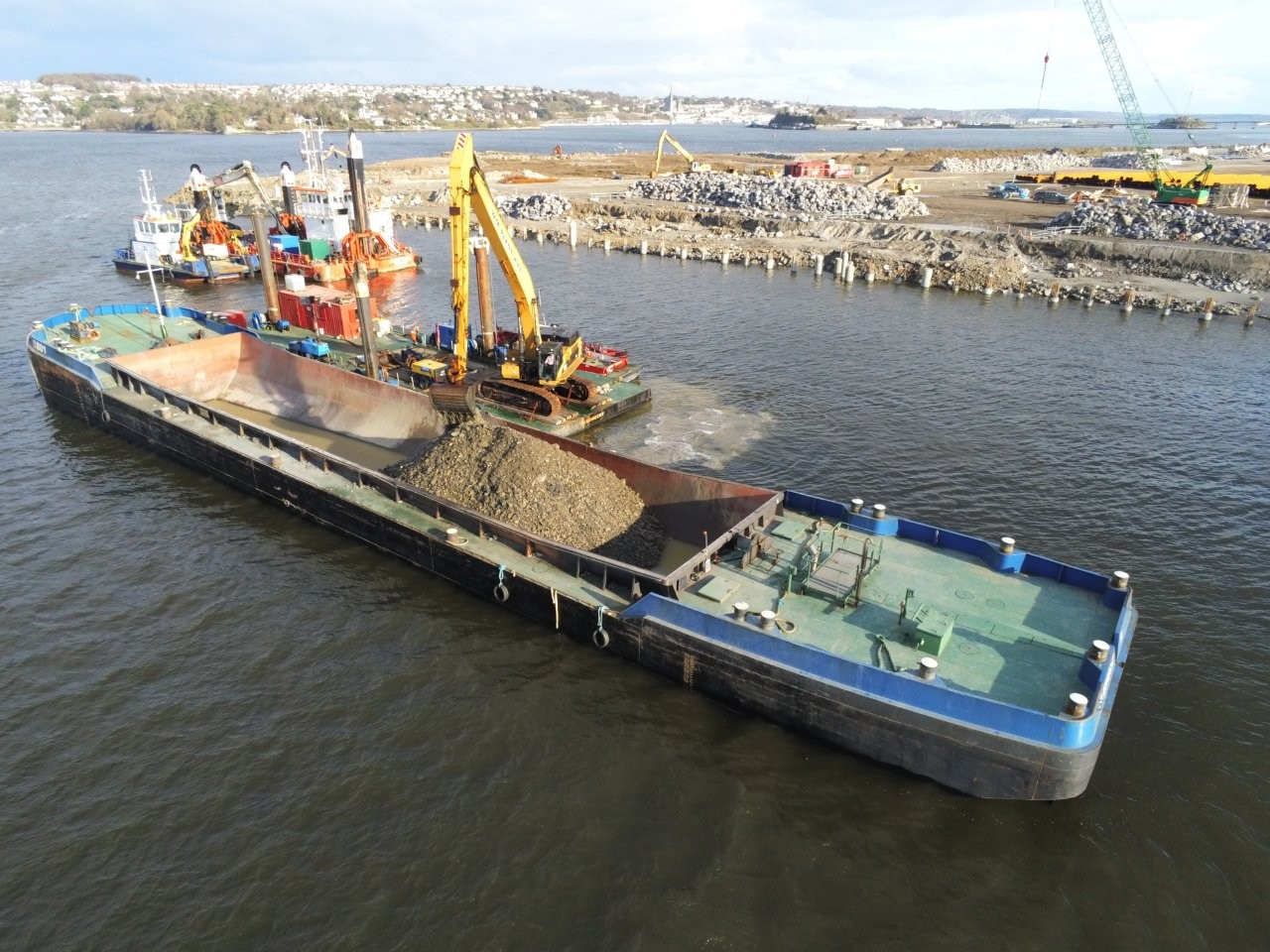 Aerial view of a barge partially filled with gravel as a large yellow excavator loads material onto it, with additional construction vessels and equipment working along a shoreline in the background beside a body of water.