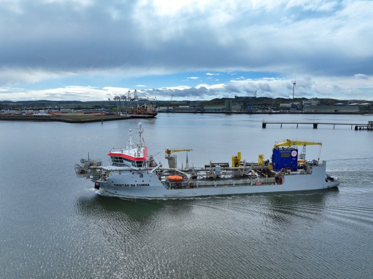 Large industrial vessel travelling through a harbour, equipped with pipes and machinery on deck, with port infrastructure, cranes, and a wind turbine visible in the background beneath a cloudy sky.